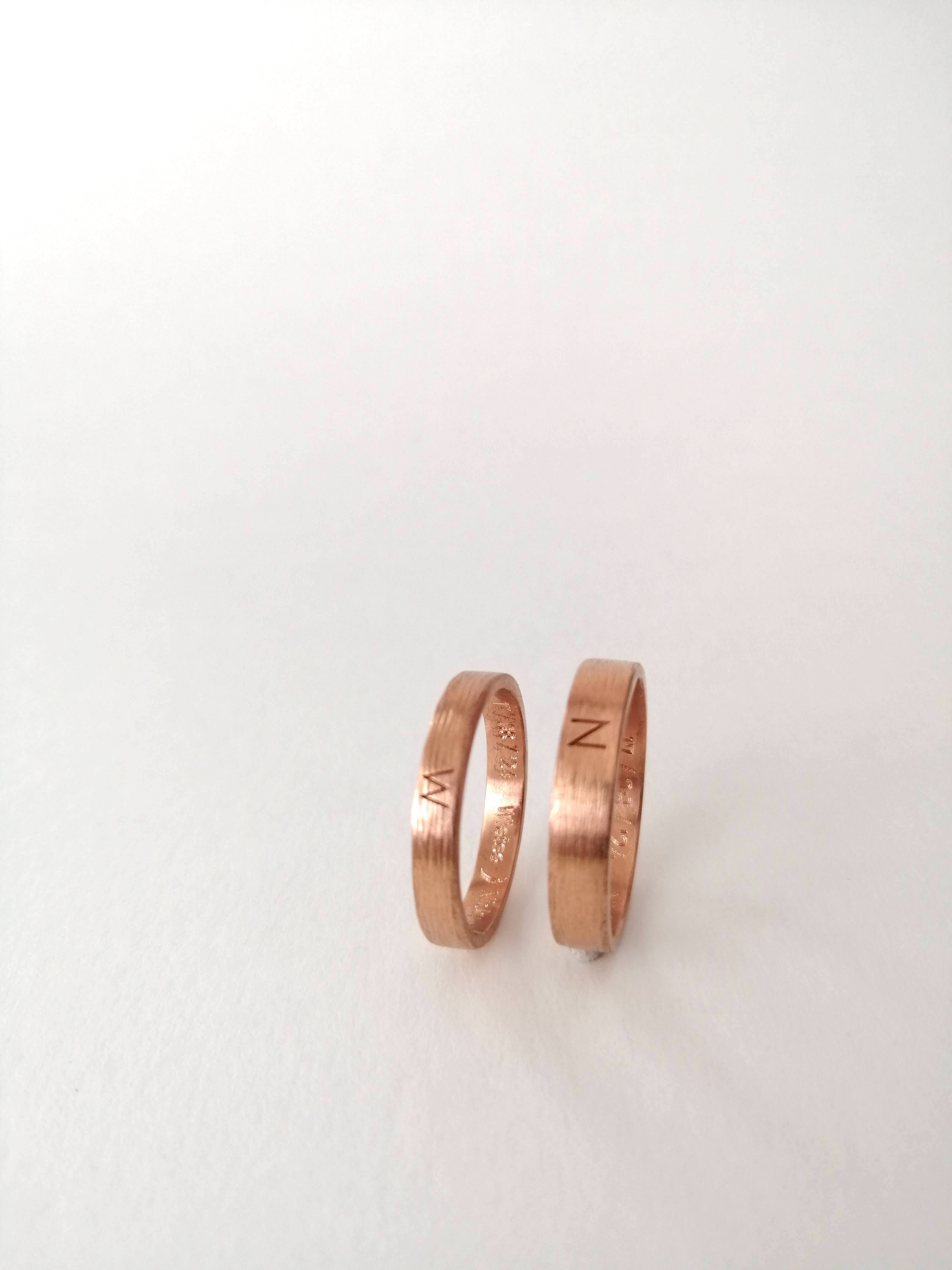 A modern wedding rings set in rose gold. Each ring is engraved with a letter on the outside, representing the spouse. The rings are a matching set, with the men's ring wider than the women's