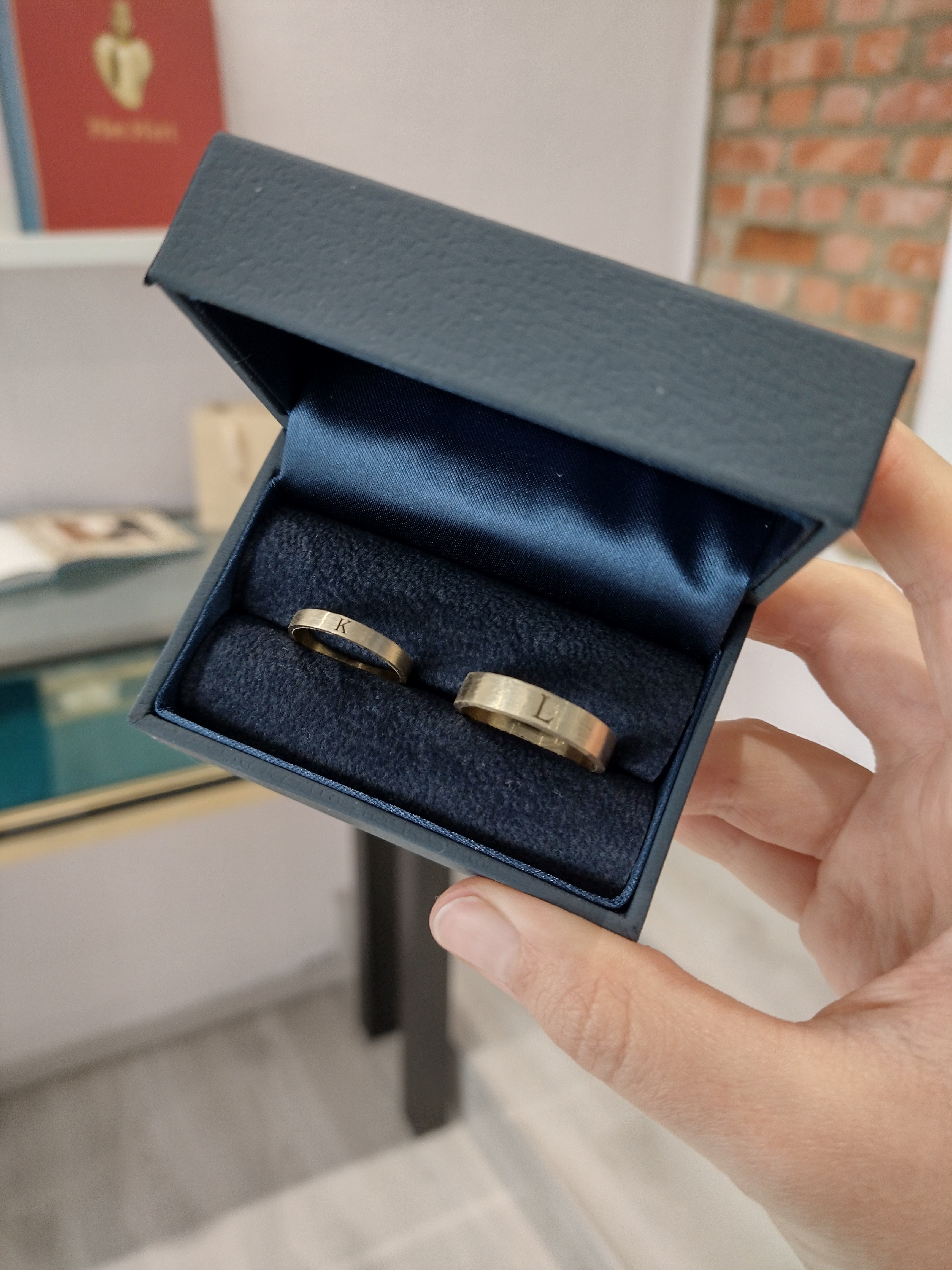 A modern wedding rings set in yellow gold. Each ring is engraved with a letter on the outside, representing the spouse. The rings are a matching set, with the men's ring wider than the women's. The rings are shown in a beautiful dark blue leatherette jewelry box.
