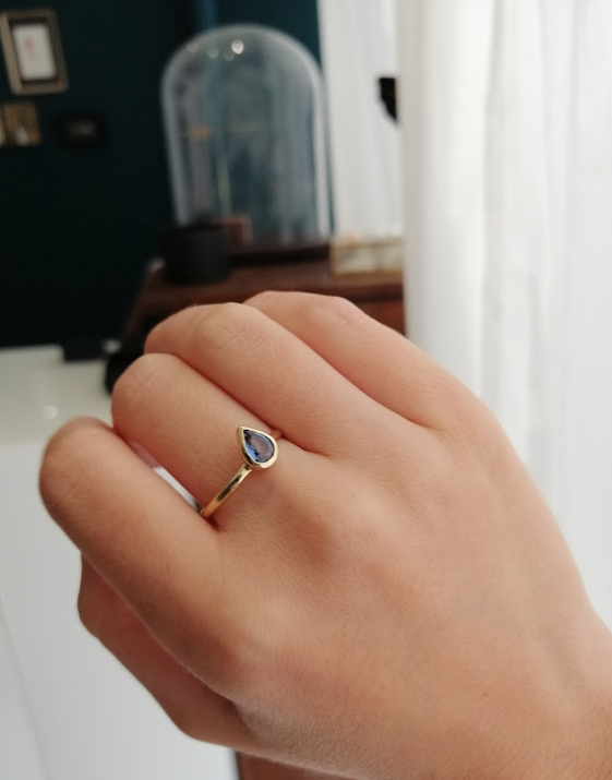 A gold engagement ring with a teardrop cut light blue sapphire set in a bezel. The ring is shown on a hand, on the middle finger