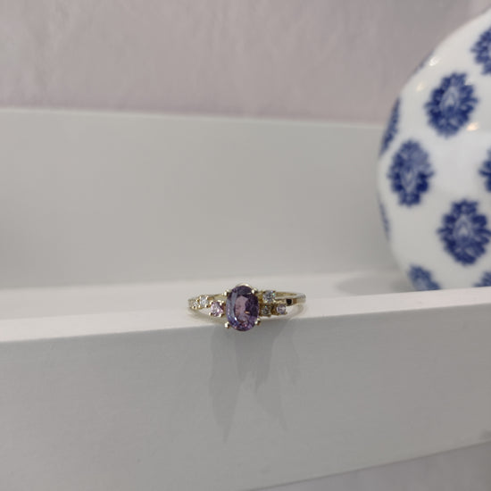 A video of a gemstone engagement ring featuring a central oval purple sapphire gemstone surrounded by a delicate cluster of smaller round gemstones in shades of purple and diamonds. The gemstones are set in a clean sleek setting with a thin gold ring band